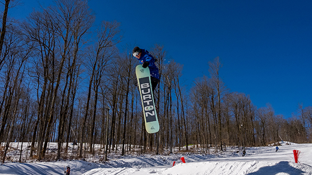 A Snowboard Instructor hits a jump and grabs his board.